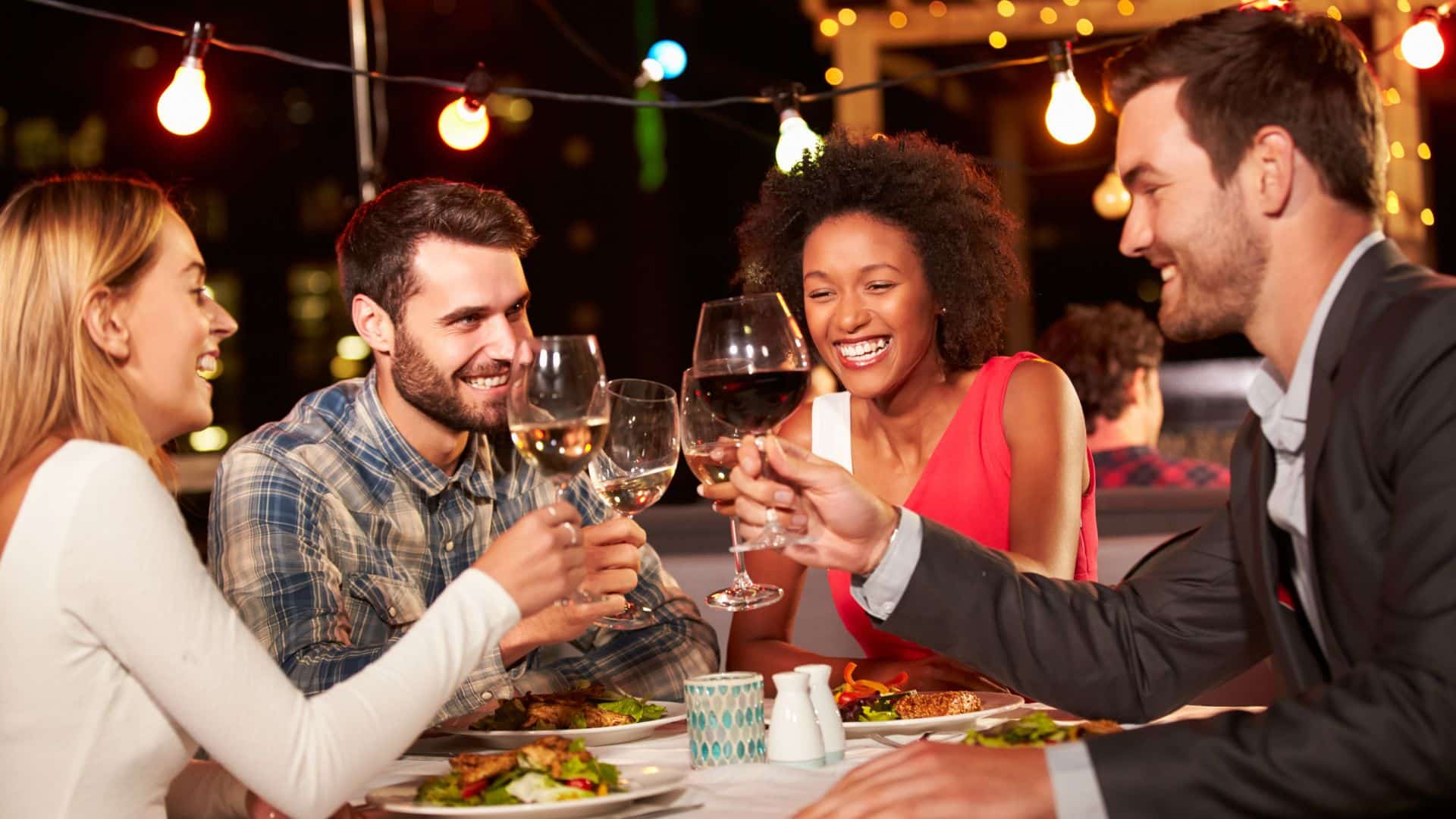 two men and two women clinking goblets while eating at a rooftop restaurant with edison lights strung in the background