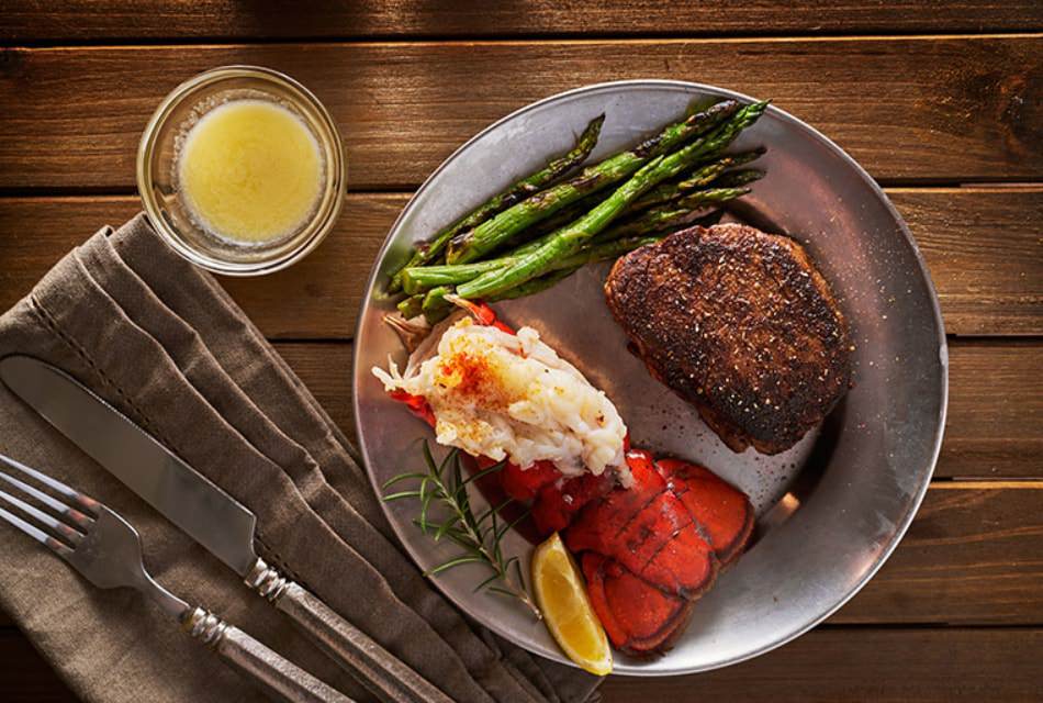 Lobster tail and filet steak with asparagus on metal plate with cutlery and ramekin of butter on wooden table.